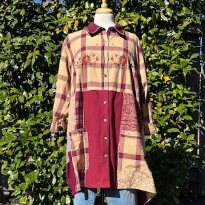 May include: A long-sleeved button-down shirt featuring a patchwork design of tan and maroon plaid fabric, solid maroon panels, and floral embroidery on the chest. The shirt includes two front pockets and a pointed collar, worn over blue jeans.