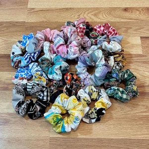 May include: A collection of 20 colorful fabric scrunchies with various patterns, including floral, plaid, and stripes. The scrunchies are arranged in a circle on a wooden surface.