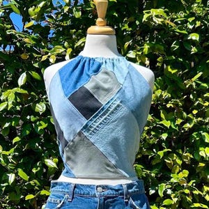 May include: A halter neck top constructed from a patchwork of denim fabric pieces in various shades of blue, grey, and black. The garment features a gathered neckline and is displayed on a mannequin torso, paired with blue denim jeans.