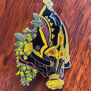 May include: A black and gold enamel pin featuring a detailed illustration of a stylized face with a floral design. The face has red eyes and a yellow crown. The flowers are yellow and green.