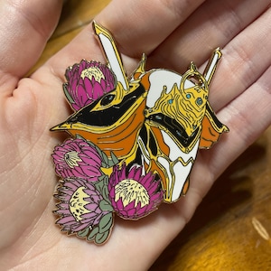 May include: A gold-toned enamel pin featuring a stylized white and orange robot head with a yellow crown and black eyes. The robot head is surrounded by pink and yellow flowers.