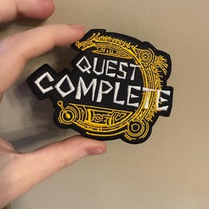 Peut inclure: Un écusson brodé noir avec un motif doré. L'écusson porte le texte "QUEST COMPLETE" en lettres blanches.