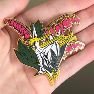 May include: Gold-tone enamel pin featuring a white and black stylized face with a crown and a floral design. The flowers are pink and green.