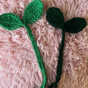 Pode incluir: Dois caules de folhas de crochê verdes sobre um fundo rosa peludo. Os caules são feitos de fio verde e têm duas folhas cada.