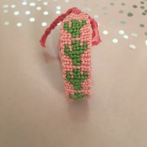 以下が含まれることがあります： 手作りのピンクとグリーンのカギ針編みブックマーク。サボテンのデザインです。ブックマークの上部には、編み込みのピンクのタッセルが付いています。サボテンの模様は緑色で、背景はピンク色です。