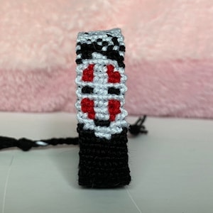 May include: Black, white, and red friendship bracelet with a stylized face design. The bracelet is made of woven string.