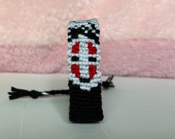 May include: Black, white, and red friendship bracelet with a stylized face design. The bracelet is made of woven string.