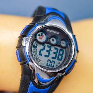 May include: A black and blue digital wristwatch with a silver trim. The watch face displays the time as 23:38 and the date as 4. The watch also has a stopwatch and alarm function. The watch is water resistant to 50 meters.