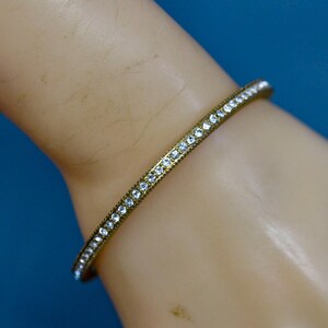 May include: A gold-coloured bangle bracelet set with small, clear crystals. The bracelet is on a light-skinned hand against a blue background. The crystals are evenly spaced along the bracelet.