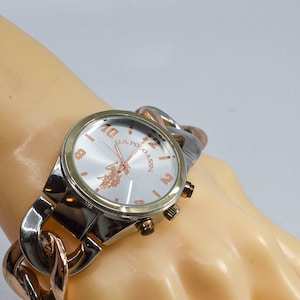 May include: A silver and rose gold watch with a white face and a chain link band. The watch face has a logo that says "U.S. POLO ASSN."