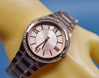 May include: A rose gold watch with a silver face and Roman numeral hour markers. The watch has a crystal-encrusted bezel and a metal band.