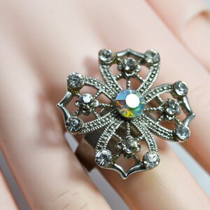 May include: A silver ring featuring a large, ornate flower design. The flower is crafted from metal and is adorned with numerous small, clear rhinestones. The centre of the flower has a larger, iridescent rhinestone.