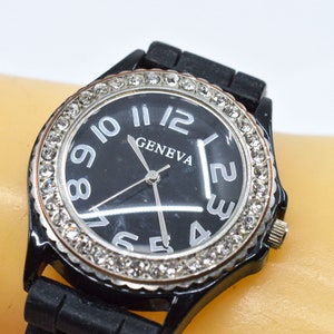 May include: Black Geneva watch with a black band and a black watch face. The watch face has silver numerals and hands, and is surrounded by a ring of clear rhinestones. The watch is on a light-colored surface.