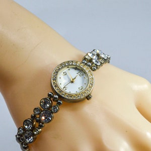 May include: A gold-tone watch with a white face and a crystal-encrusted band. The watch has Roman numeral hour markers and a second hand.