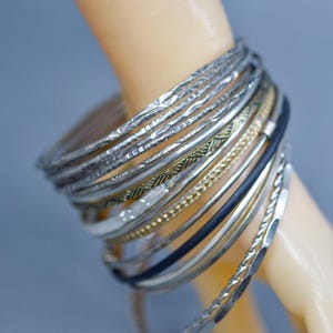 May include: A collection of silver and gold bangles on a wrist. The bangles have various textures, including hammered, twisted, and smooth designs. Some bangles are black or gold. The jewelry is stacked together, creating a layered look.