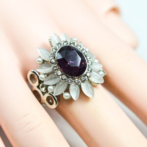 May include: A silver ring featuring a large purple gemstone surrounded by smaller white gemstones and white flower-shaped accents.