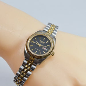 Könnte beinhalten: Eine zweifarbige Gold- und Silber-Armbanduhr mit schwarzem Zifferblatt und Datumsanzeige. Auf dem Zifferblatt der Uhr steht "SHARP" gedruckt.