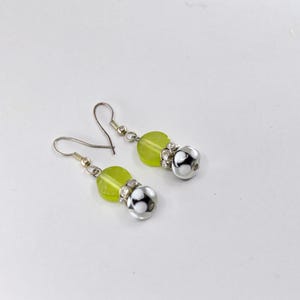 May include: A pair of drop earrings with silver hooks. Each earring has a light green, translucent bead, a silver bead with clear rhinestones, and a silver bead with a black and white pattern. The earrings are set against a white background.
