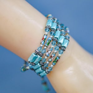 May include: A multi-strand bracelet with turquoise and silver beads. The bracelet features rectangular turquoise beads, small silver beads, and decorative enamel beads with floral designs. The bracelet is wrapped around a wrist.