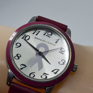 Peut inclure: Une montre argentée avec un bracelet rose et un cadran blanc. Le cadran comporte des chiffres et des aiguilles noirs, ainsi qu'un ruban rose au centre. Le cadran de la montre indique "Dana Buchman".
