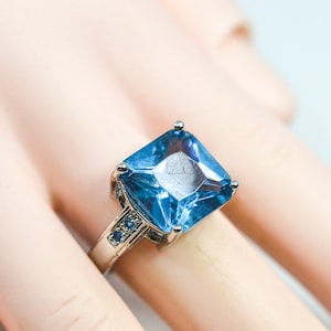 May include: A silver ring featuring a large, square-shaped blue gemstone. The gemstone is surrounded by smaller blue gemstones.