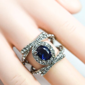 May include: A silver ring featuring a large blue oval gemstone surrounded by smaller clear gemstones. The ring has a criss-cross design.