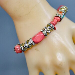May include: A pink beaded bracelet with silver-tone accents and decorative beads. The bracelet features a mix of round and rectangular pink beads, interspersed with silver-tone rings and decorative floral beads. The bracelet is displayed on a light-toned hand.