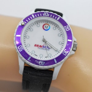Könnte beinhalten: Weiße Armbanduhr mit einer violetten Lünette und einem schwarzen Band. Das Zifferblatt der Uhr hat einen weißen Hintergrund mit schwarzen Zeigern und einem roten Logo mit der Aufschrift "Bravecto/Pluralaner".