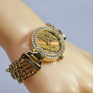 May include: Gold-tone watch with a chain link strap and a round face featuring a sparkly gold and black zebra print design. The face is surrounded by clear rhinestones.