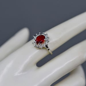 May include: A silver ring with a large oval, deep red gemstone, surrounded by smaller clear stones. The ring is displayed on a white hand against a gray background. The ring's design is classic and elegant.