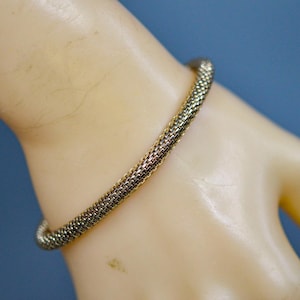 May include: A gold-toned mesh bracelet. The bracelet is a continuous loop, with a woven texture. The bracelet is worn on a light-skinned arm, with a blue background.