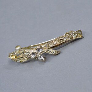 May include: A silver-coloured barrette with a floral design. The barrette features clear, sparkling rhinestones and intricate detailing. The clip is on a grey background.