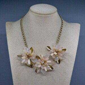 May include: A statement necklace featuring three floral clusters made of translucent pale pink and white petals with gold-toned metallic leaves. The floral elements are connected by a gold-toned chain that rests around the neck area.