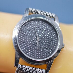May include: Silver-toned wristwatch with a round face covered in small, sparkling embellishments. The watch has a matching silver band with a textured pattern. The watch is shown on a wrist.
