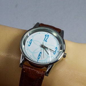 Könnte beinhalten: Eine silberne Armbanduhr mit einem braunen Lederarmband. Das Zifferblatt der Uhr hat einen weißen Hintergrund mit einem blauen Blumendesign und blauen Zahlen.