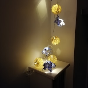 Peut inclure: Une guirlande de lanternes en papier avec un motif géométrique bleu et blanc et un motif géométrique jaune et blanc. Les lanternes sont allumées et suspendues à un cordon blanc.
