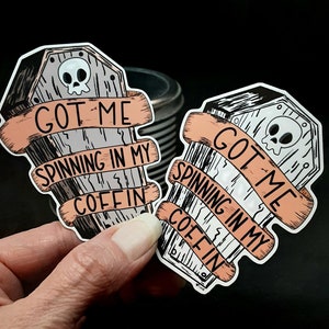 May include: Two black and white coffin-shaped stickers with a skull and crossbones on each. The text on the stickers reads "Got me spinning in my coffin".