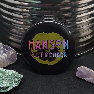 May include: A black button with the text "HANSON CULT MEMBER" in rainbow colors. The button is surrounded by several small, clear and purple crystals on a silver metal container.