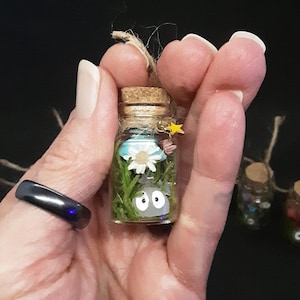 May include: A small glass bottle with a cork top filled with moss, a white flower, and a small gray figure with eyes. The bottle is decorated with a small yellow star.