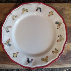 May include: White ceramic plate with a scalloped edge and a red border. The plate is decorated with colorful rooster illustrations around the rim. The plate is on a wooden surface.