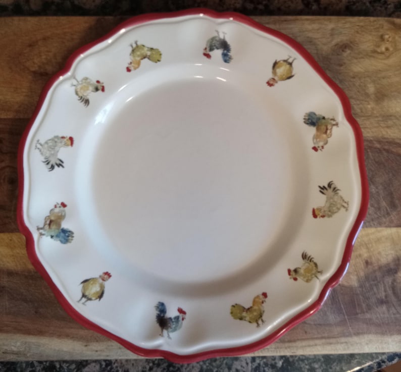 May include: White ceramic plate with a scalloped edge and a red border. The plate is decorated with colorful rooster illustrations around the rim. The plate is on a wooden surface.