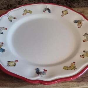 May include: White ceramic plate with a scalloped edge and a red border. The plate is decorated with colorful rooster illustrations around the rim. The plate is round and has a smooth surface.