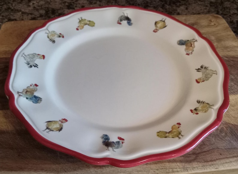 May include: White ceramic plate with a scalloped edge and a red border. The plate is decorated with colorful rooster illustrations around the rim. The plate is round and has a smooth surface.