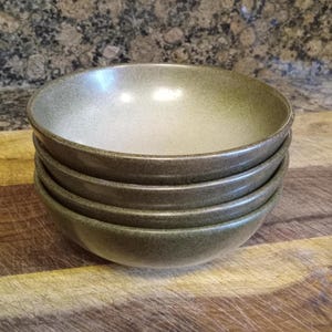 May include: A stack of four round ceramic bowls featuring a speckled olive green exterior and a smooth, light beige interior. The bowls have a simple, wide-rimmed design with a consistent shape, stacked vertically to display their uniform size and color.