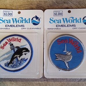 May include: Two circular Sea World embroidered emblems on white backing cards. The left patch features a black orca jumping near a lighthouse with red text. The right patch shows a grey shark and a sailboat with red text on a blue background.