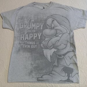 May include: A heather gray short-sleeve t-shirt featuring a graphic of the character Grumpy with arms crossed. The text reads: I'm GRUMPY You're HAPPY THINGS = EVEN OUT. The design is printed in a darker shade of gray with a distressed texture.