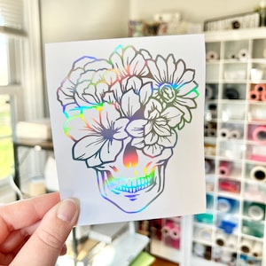 Puede incluir: Una pegatina hologr&aacute;fica con un dise&ntilde;o de calavera y elementos florales. La calavera est&aacute; adornada con varias flores. La pegatina tiene un acabado iridiscente reflectante, creando un efecto arco&iacute;ris.