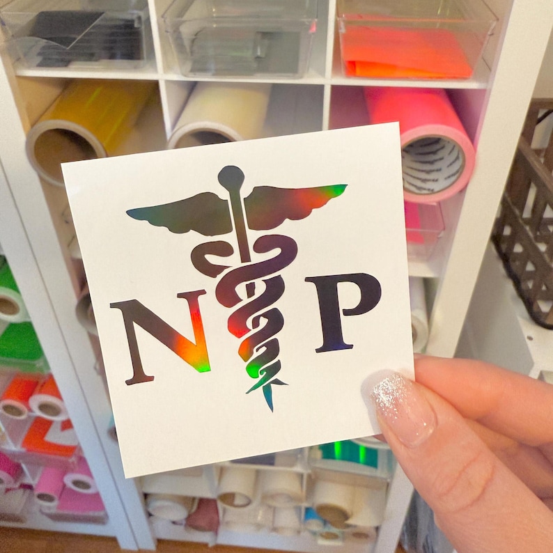 May include: A square white decal featuring a holographic caduceus symbol centered between the letters N and P. The caduceus design displays a metallic, rainbow-like color shift, while the letters N and P are rendered in a solid dark tone.
