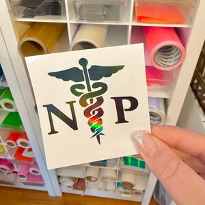 May include: A square decal featuring a holographic caduceus symbol centered between the letters N and P. The design displays a spectrum of iridescent colors that shift depending on the light, set against a plain white square backing material.