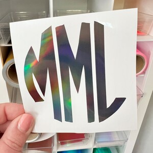 May include: A vinyl decal featuring the letters MML in a stylized, bold font. The letters have a holographic finish that reflects a spectrum of colors including green, yellow, and red against a dark, metallic base on a white backing sheet.